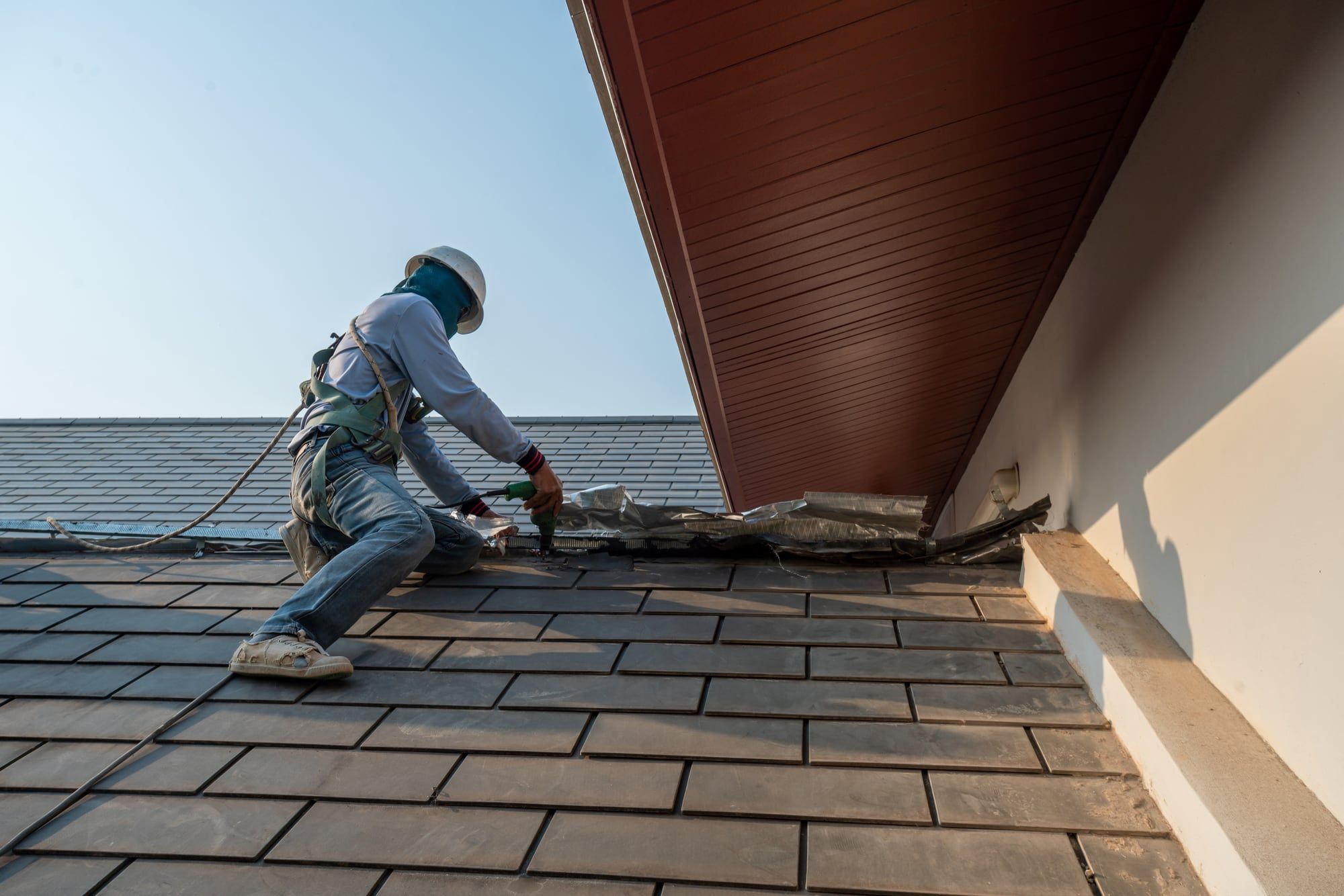 Roofer in a safety harness repairing the flashing where a shingle roof meets a wall